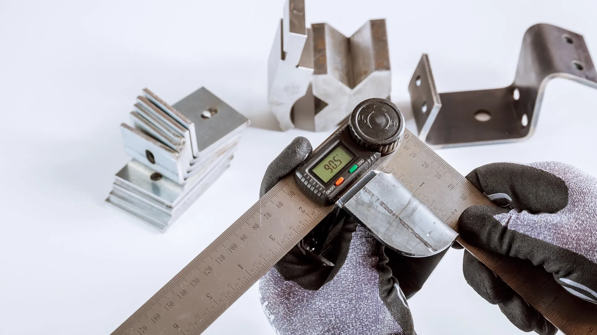 Hand using a digital angle gauge and steel ruler to check a bent sheet metal bracket next to other cut parts
