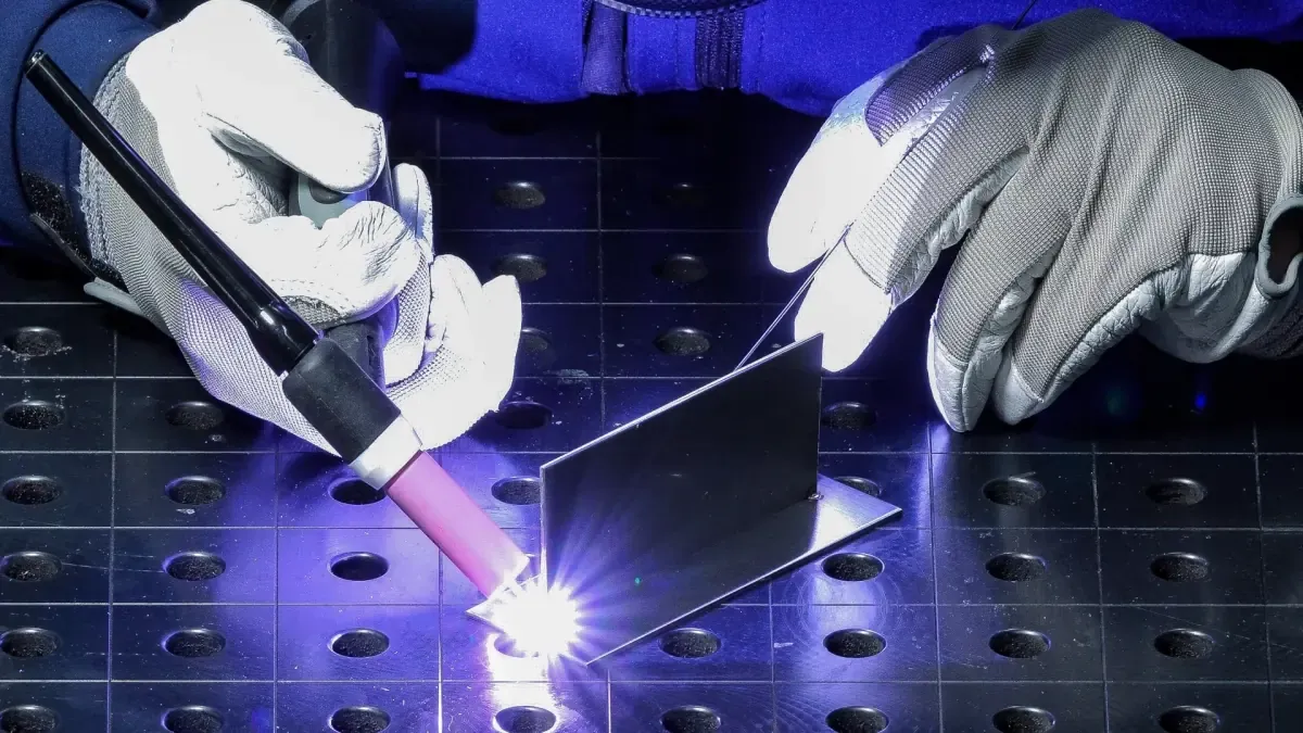 Gloved hands TIG welding a sheet metal bracket with a laser cutter cutting bed in the background