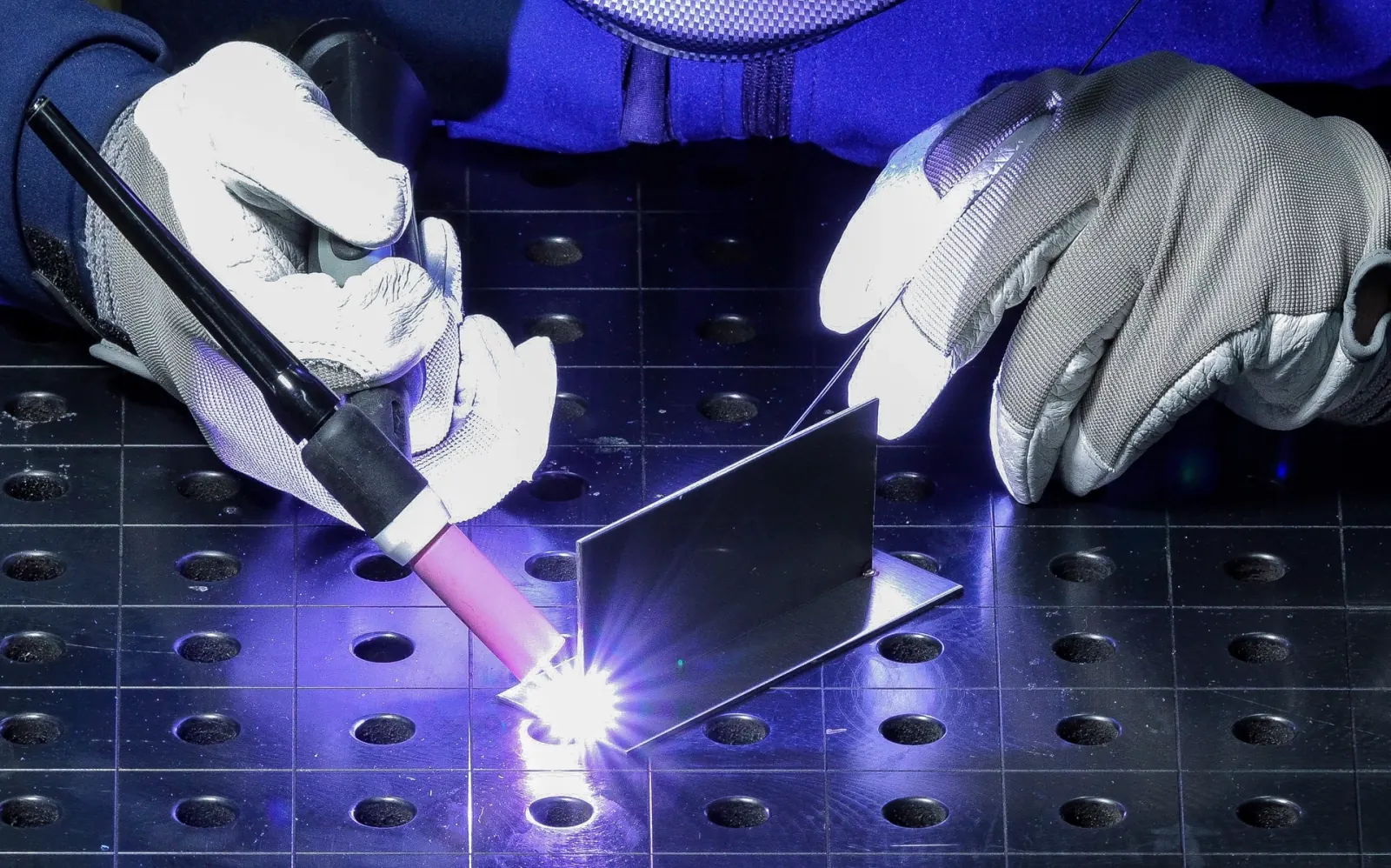 Gloved hands TIG welding a sheet metal corner bracket with bright arc light on a welding table