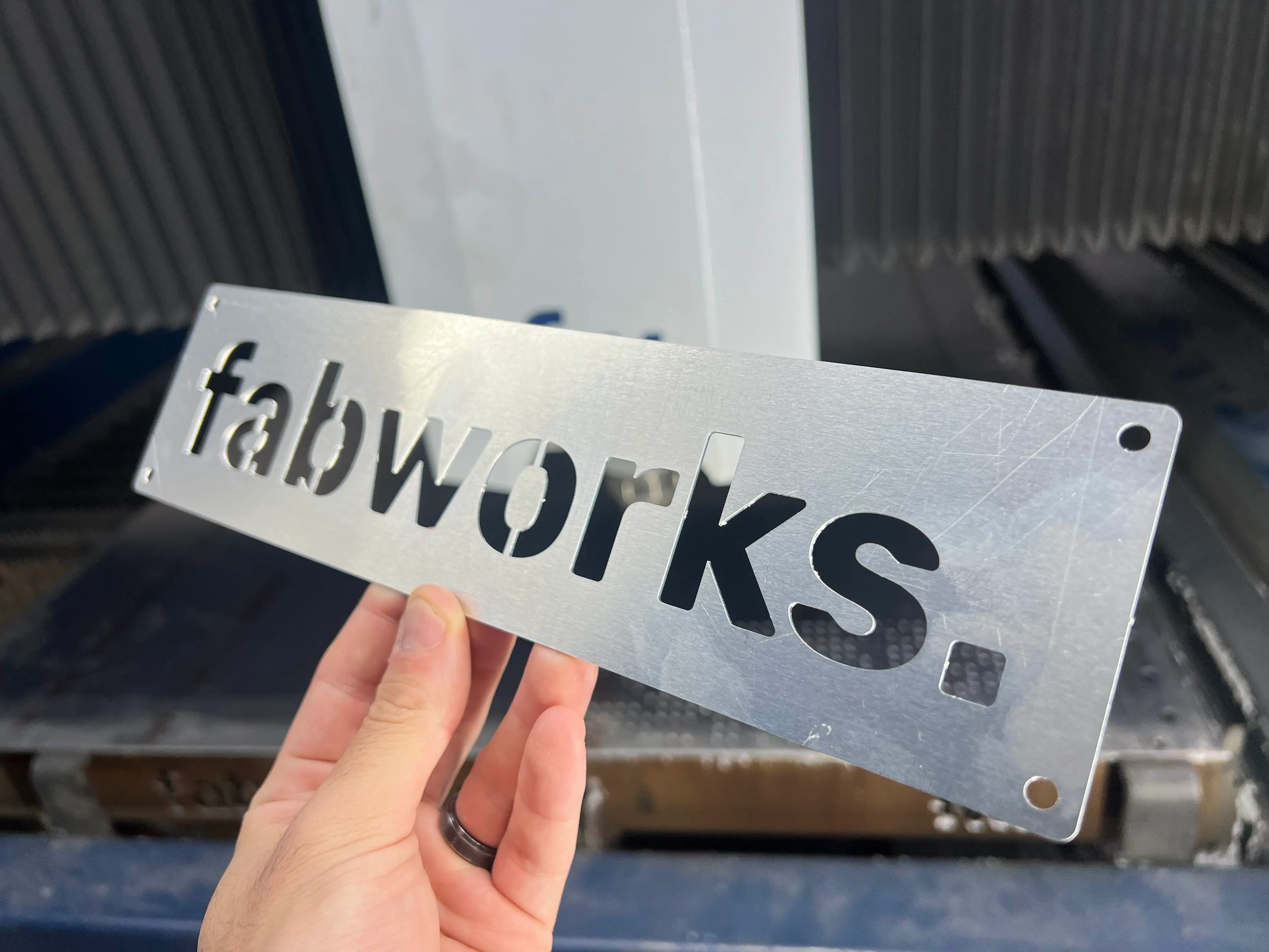 Hand holding the finished laser-cut steel fabworks sign in front of the cutting machine